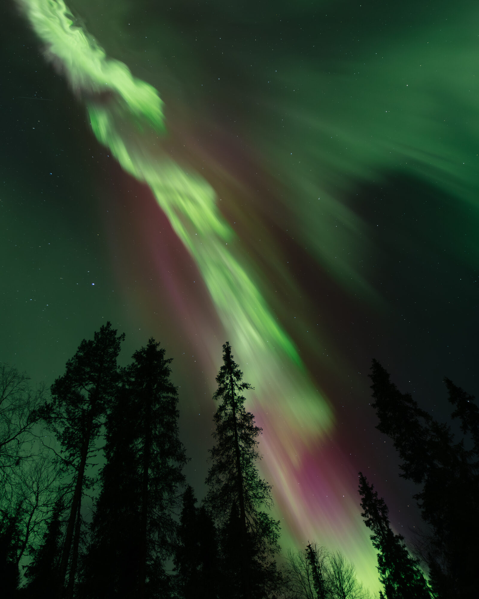 The glowing triangular window of an A-frame forest cabin beneath a green aurora streak and a sky full of stars at Sixty Eight North.