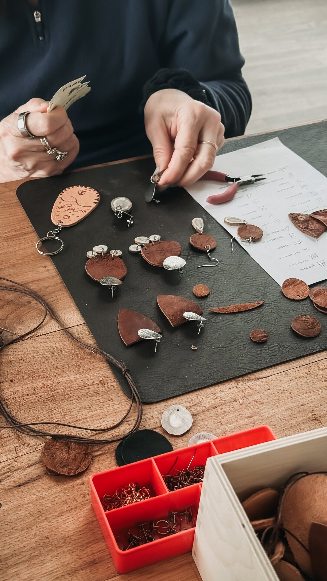 Hands at work on a wooden table laid with leather and reindeer-antler jewellery pieces, pliers and findings nearby.