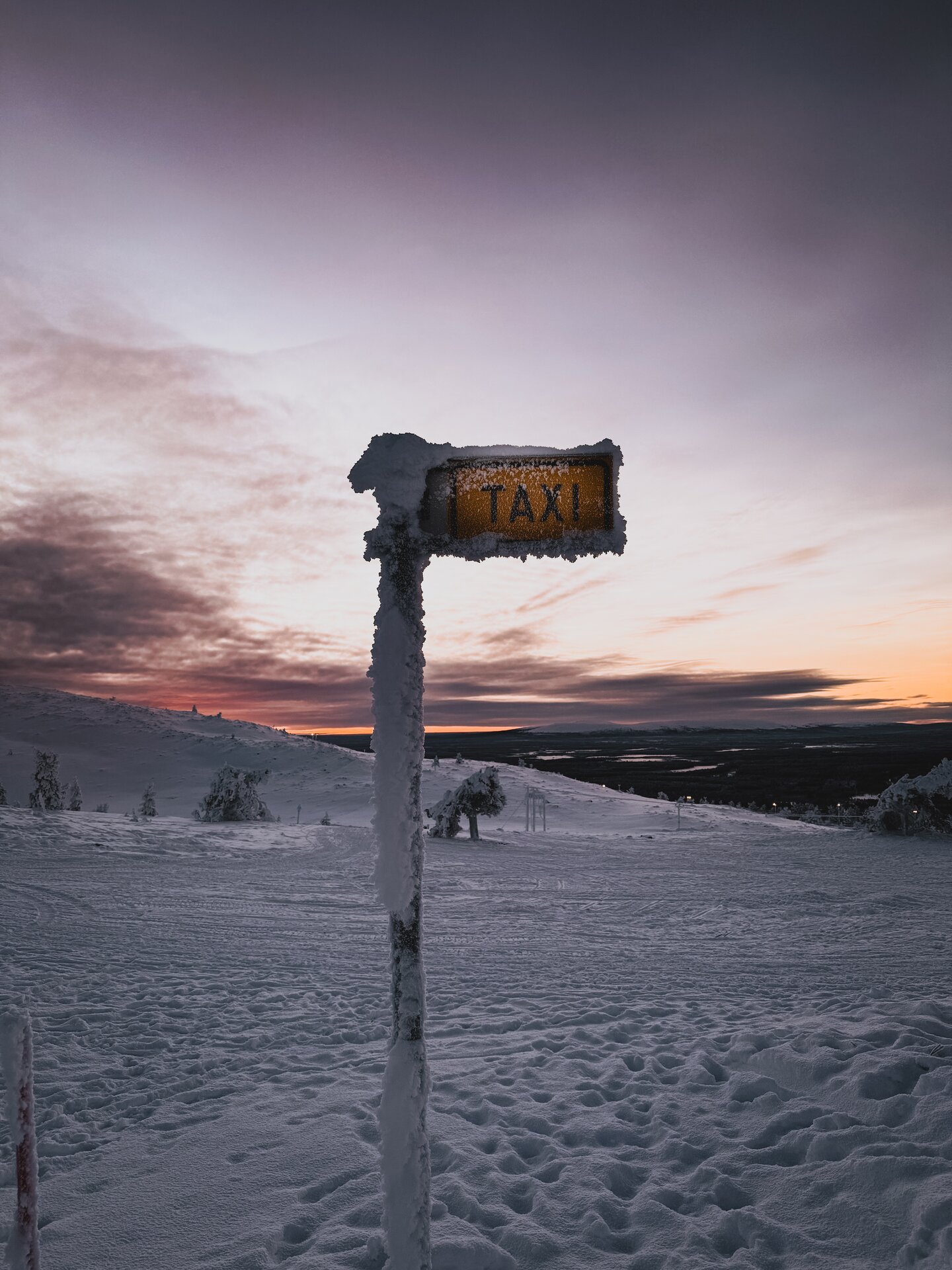 A roadside taxi sign in dusk light during winter in Finnish Lapland, snow on the ground and soft polar light in the distance.