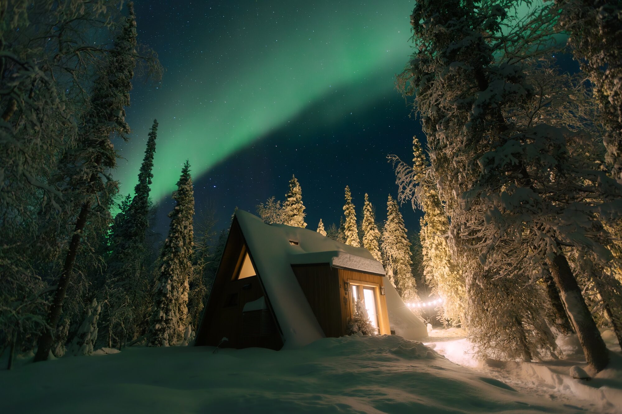 The Northern Lights arcing in a green band over a snow-covered A-frame cabin and pine forest at Sixty Eight North, near Levi in Finnish Lapland.
