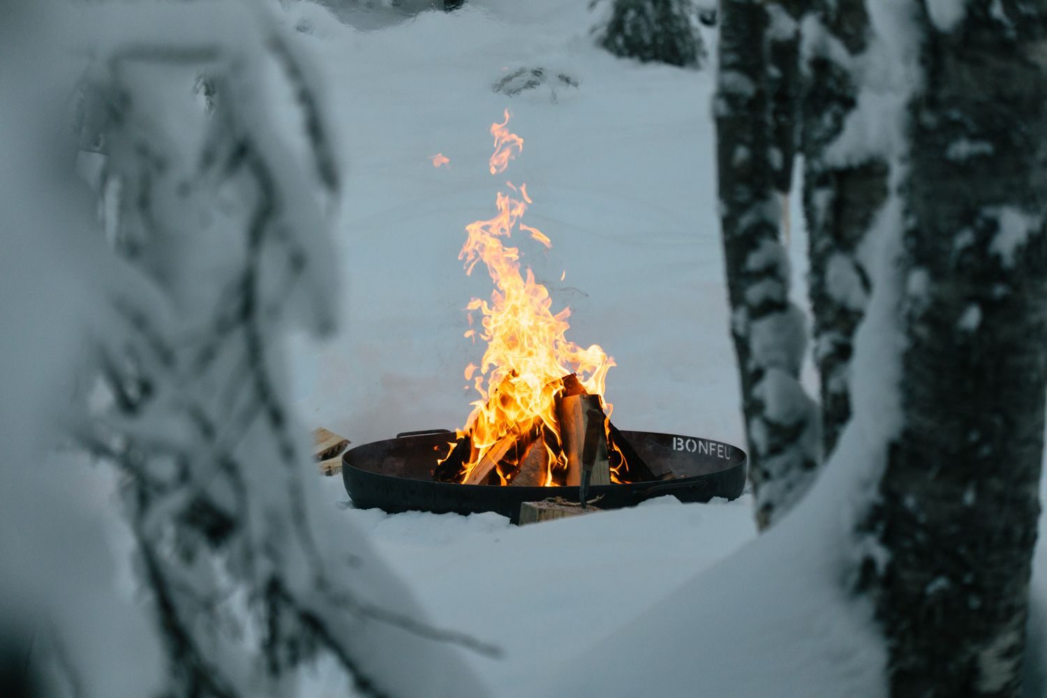 An open fire glowing warm in the winter forest at Sixty Eight North in Finnish Lapland, deep snow around the fire ring and pines silhouetted in the dark beyond.