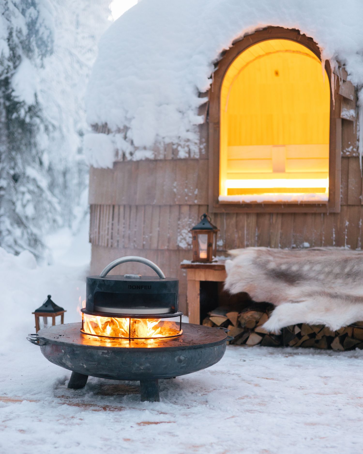 The outdoor fireplace burning beside a wooden cabin, a lantern glowing and a reindeer skin folded over the bench.