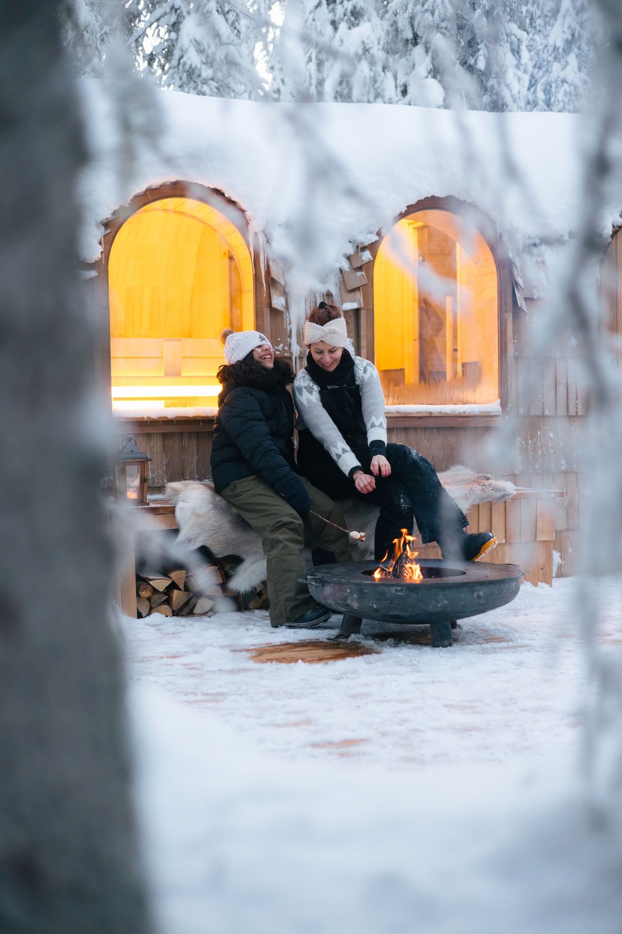 Two guests on a fur-draped bench by the outdoor fire bowl, roasting marshmallows, the warm-lit arched window behind them.