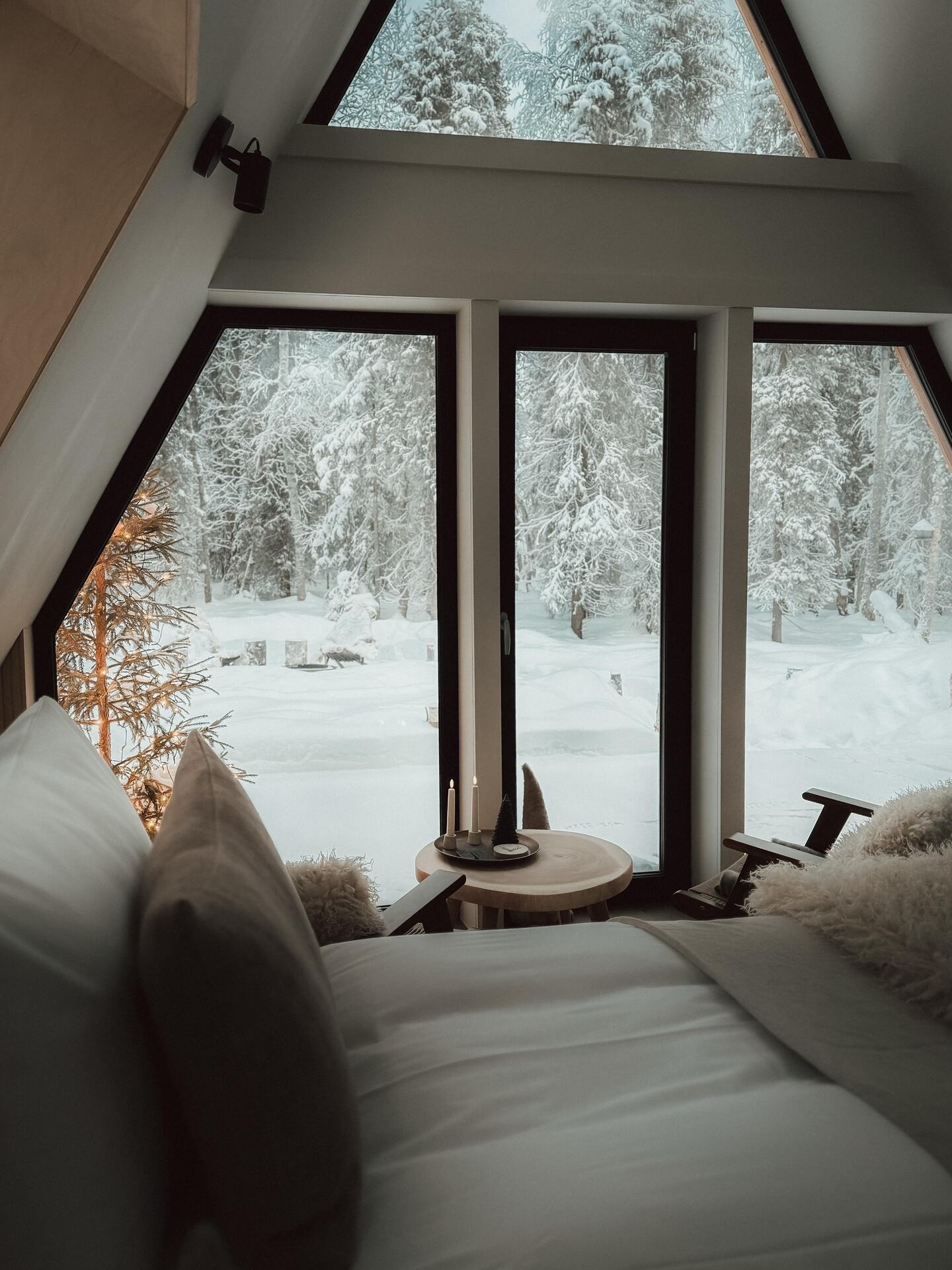 The view from the bed inside the A-frame cabin: a candlelit table and sheepskin-draped chairs at the gable windows, snow-heavy pines filling every pane.