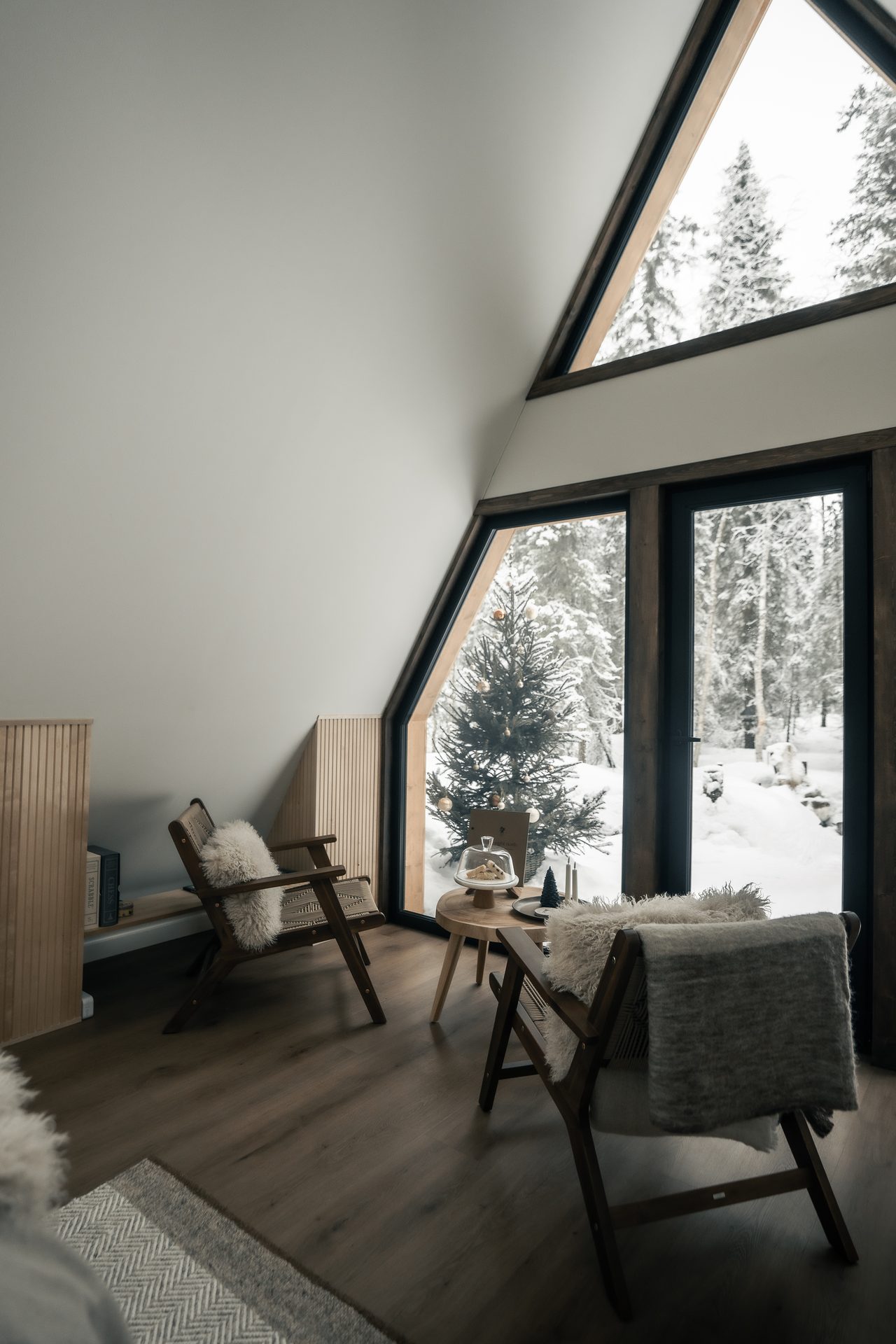 The interior of a Scandinavian A-frame cabin at Sixty Eight North, with a floor-to-ceiling triangular window looking onto snow-covered pine forest.