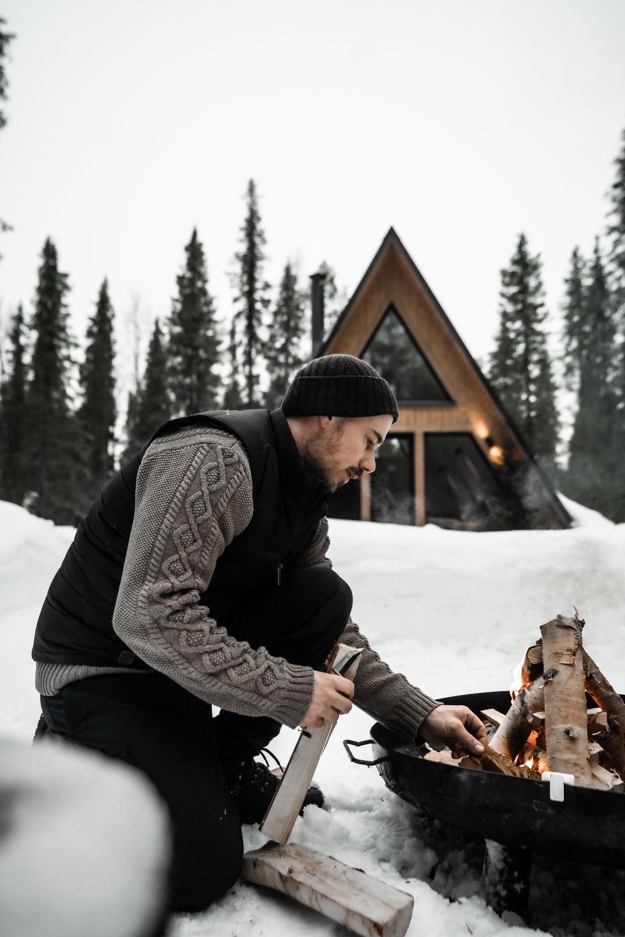 A guest tending the fire pit outside the cabin, birch logs ready, the forest cabin held in pines beyond.