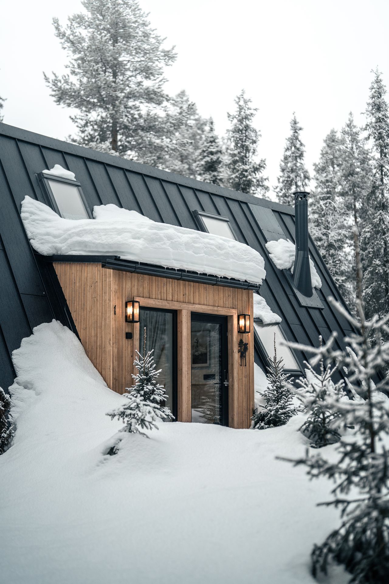 The entrance of an A-frame cabin at Sixty Eight North beneath a snow-laden roof, lanterns lit on either side of the door and small spruce saplings dressed in snow.