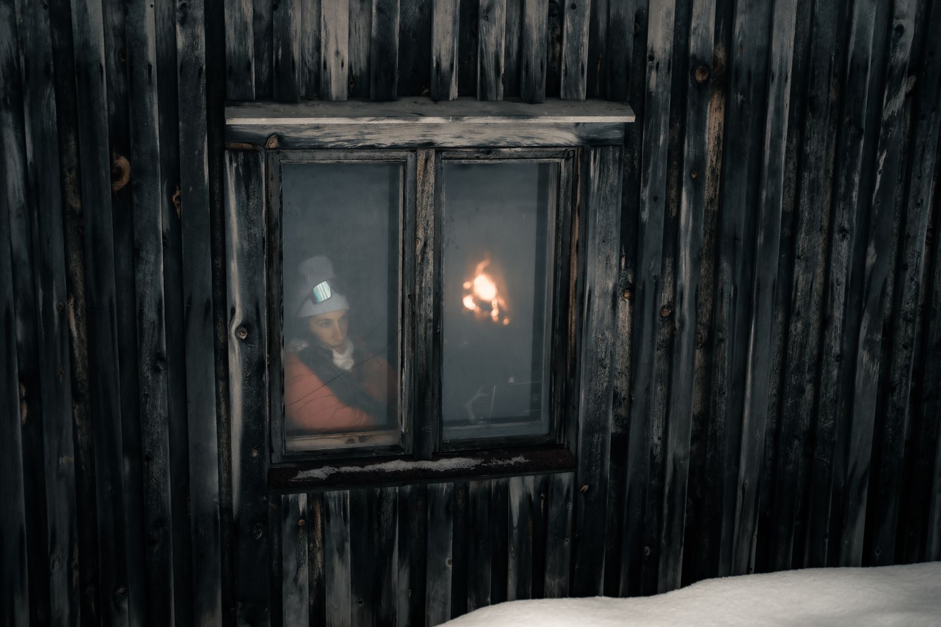 A guest seen through the window of a weathered wooden cabin, firelight warm against the dark timber walls.