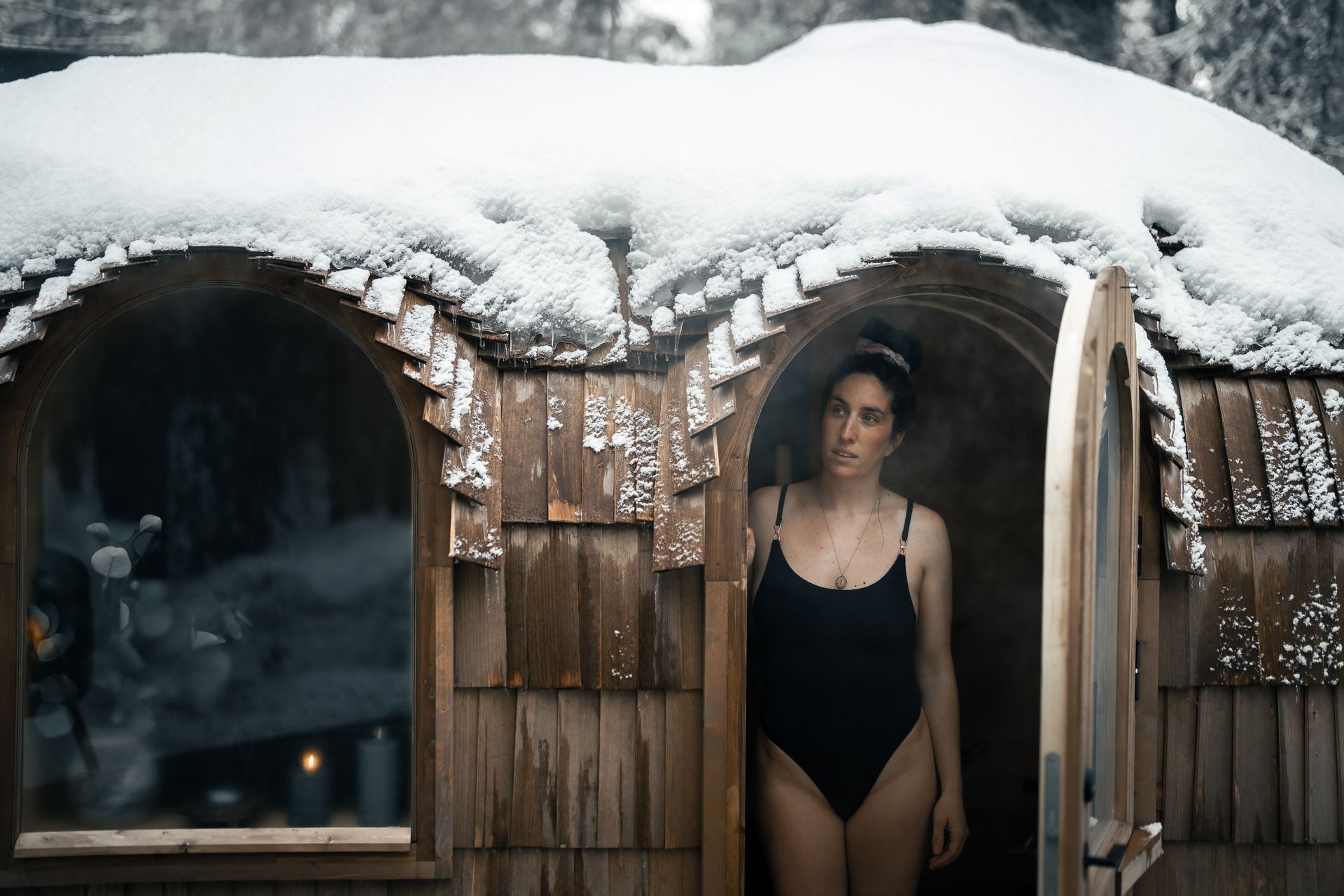A guest standing in the doorway of the barrel sauna, heavy snow on the curved roof.