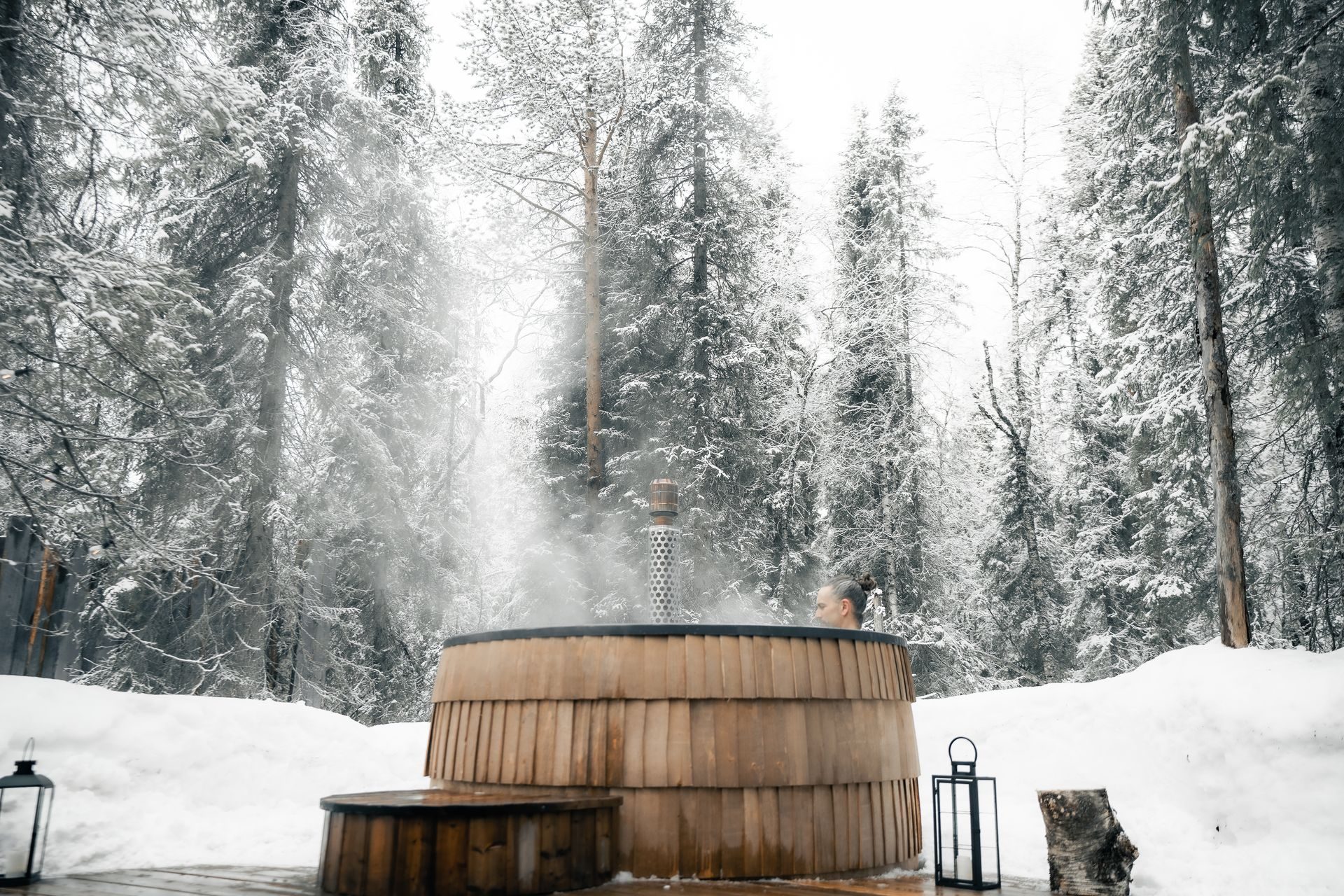 Steam rising from the round wooden hot tub at the edge of the snow-laden forest, a lantern set on the deck.