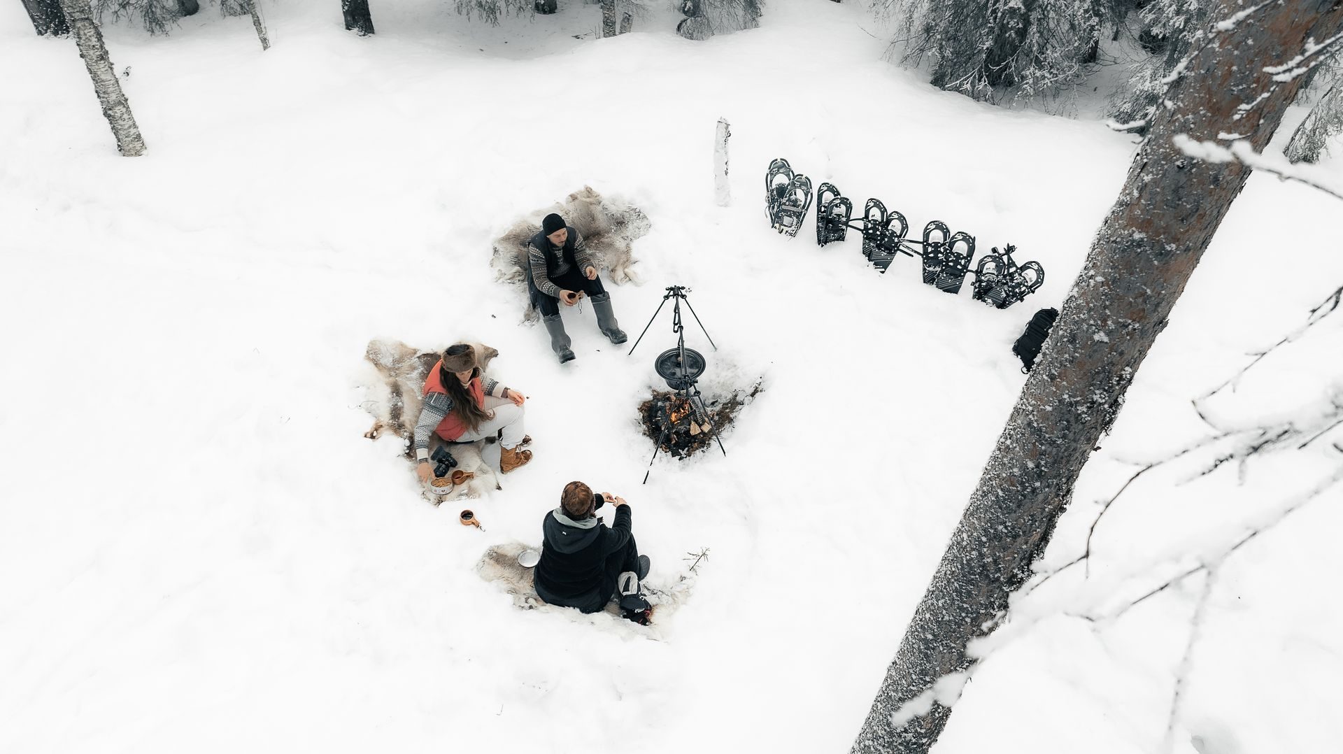 Three guests sitting on reindeer hides around an open fire in the snow with a tripod cooking pot, snowshoes lined up beside them, snowy birch and pine forest around.