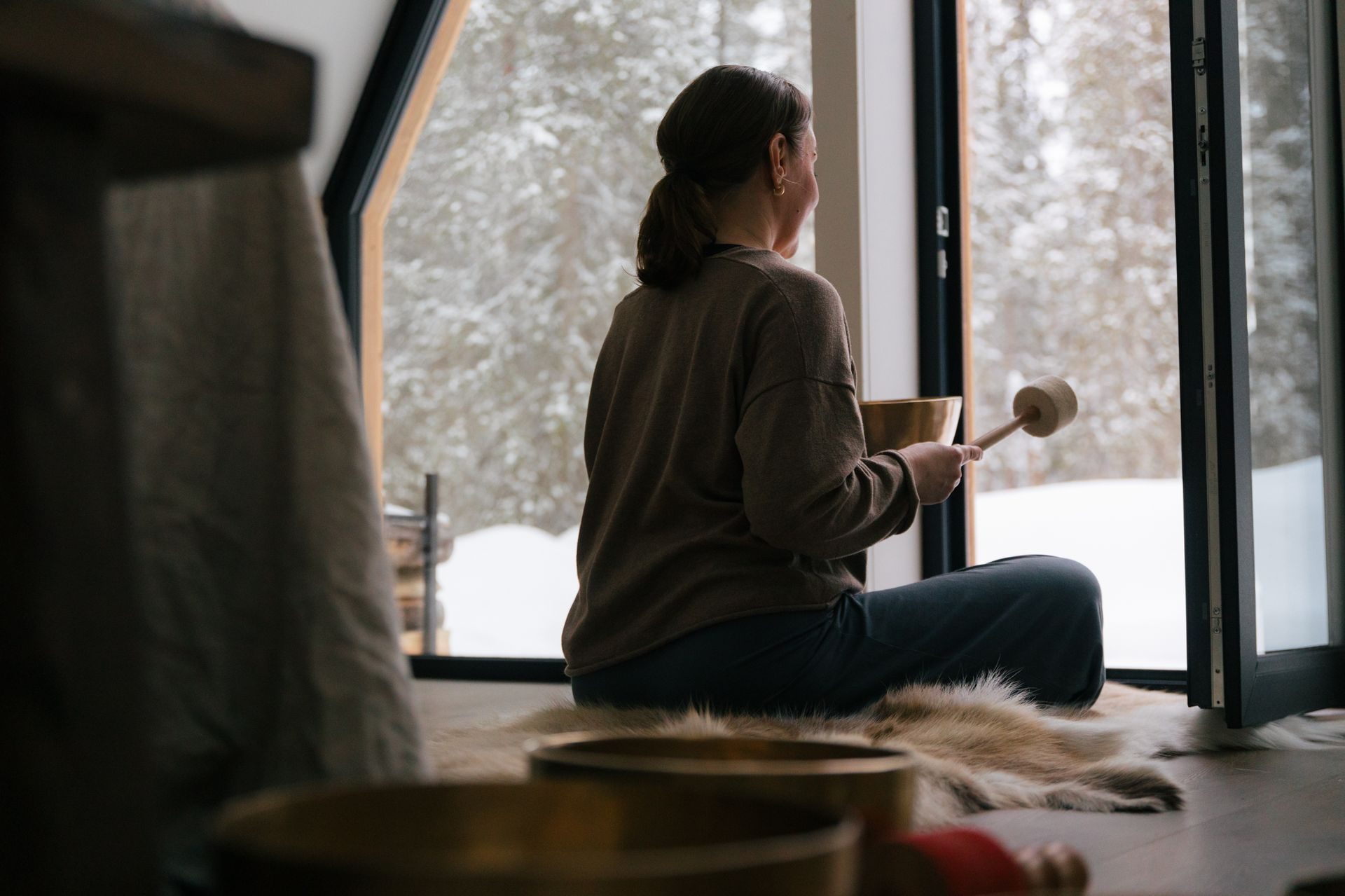 The practitioner from behind, seated on a reindeer hide by the cabin's wide window, singing bowl in hand and the snowy forest beyond.