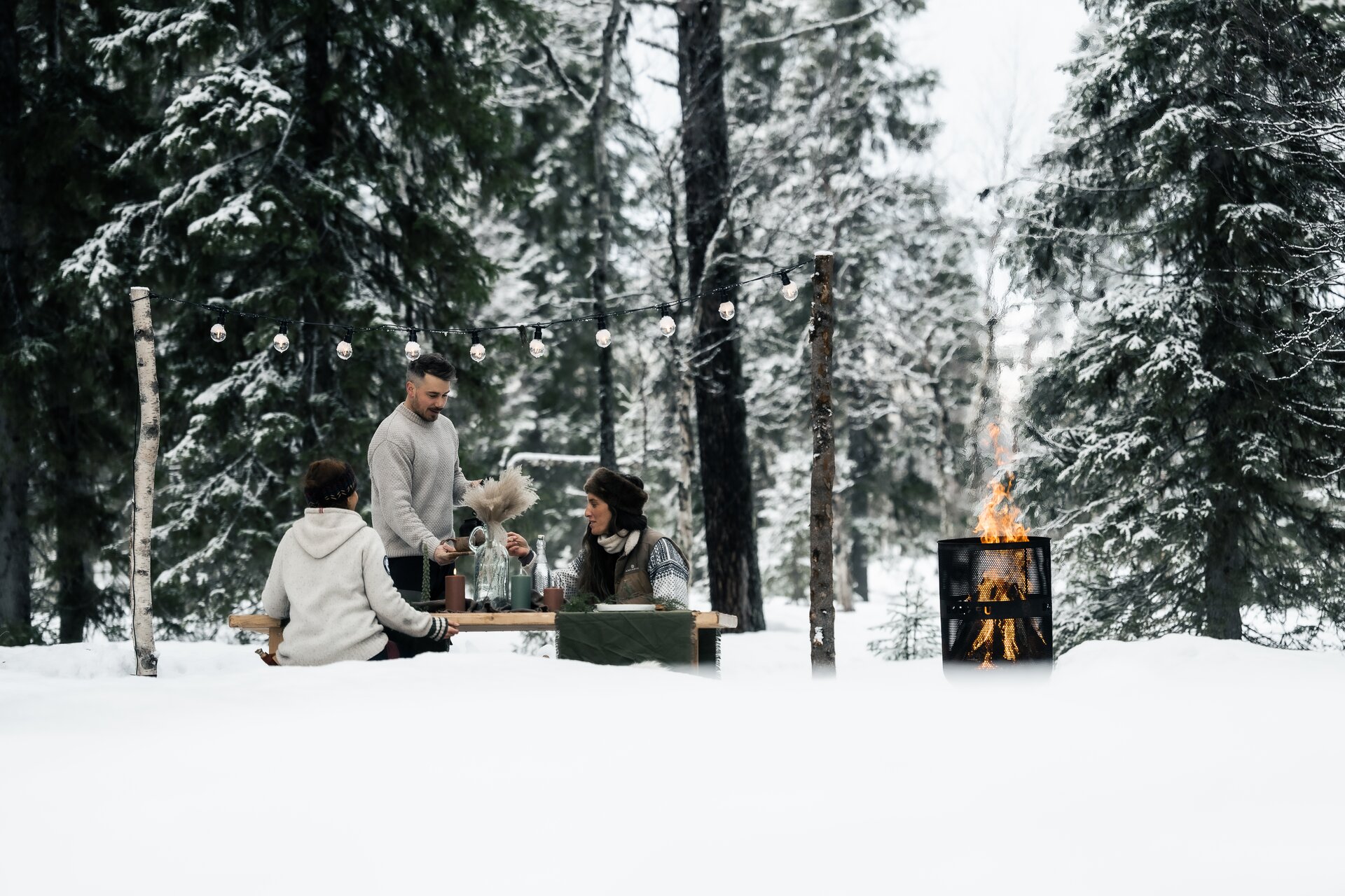 Guests gathered at a forest table with string lights and an open fire in the snow at Sixty Eight North, Finnish Lapland.