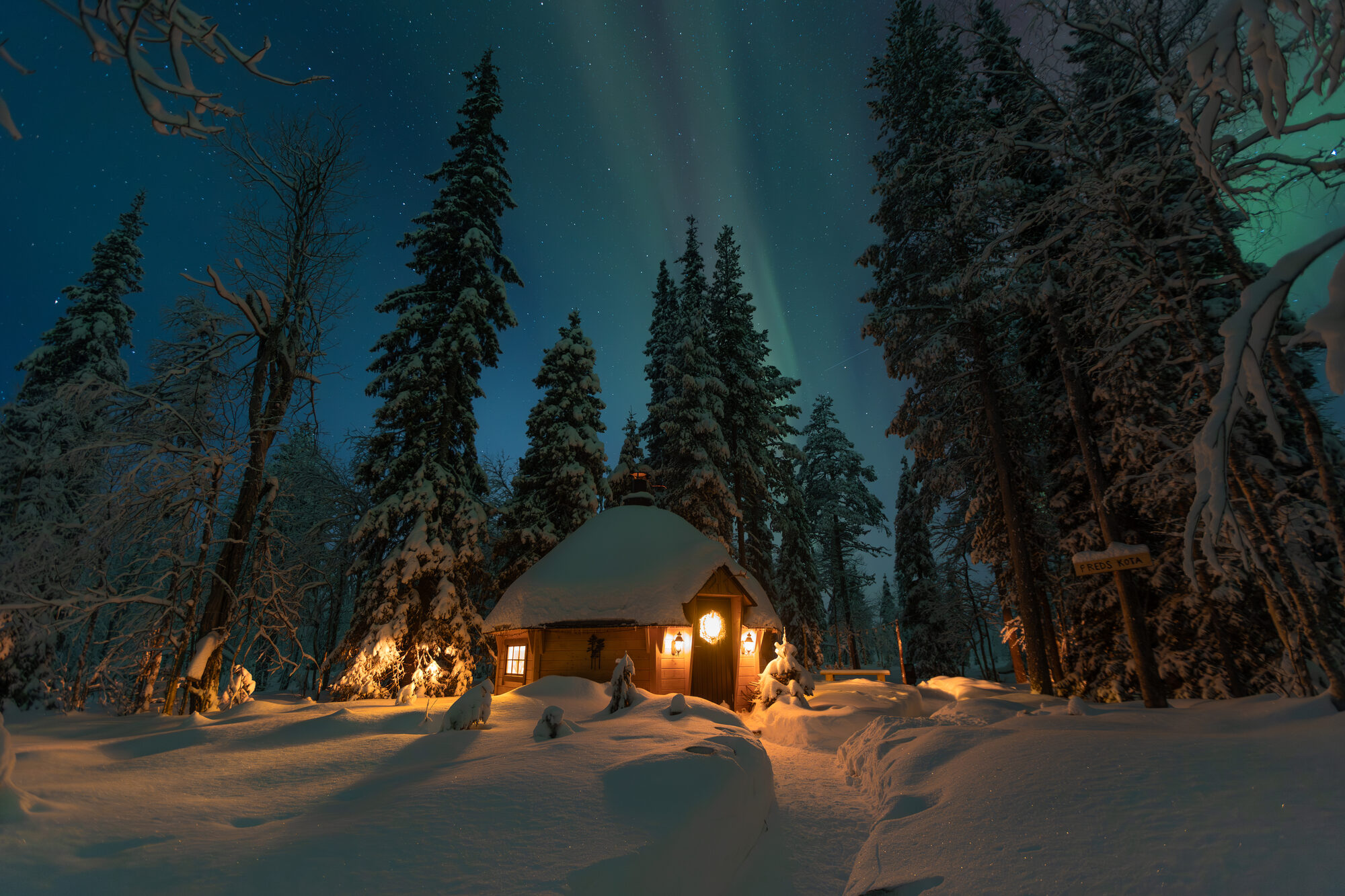 A snow-covered kota cabin glowing warmly among tall pines under a faint green aurora in the forest near Levi, Finnish Lapland.