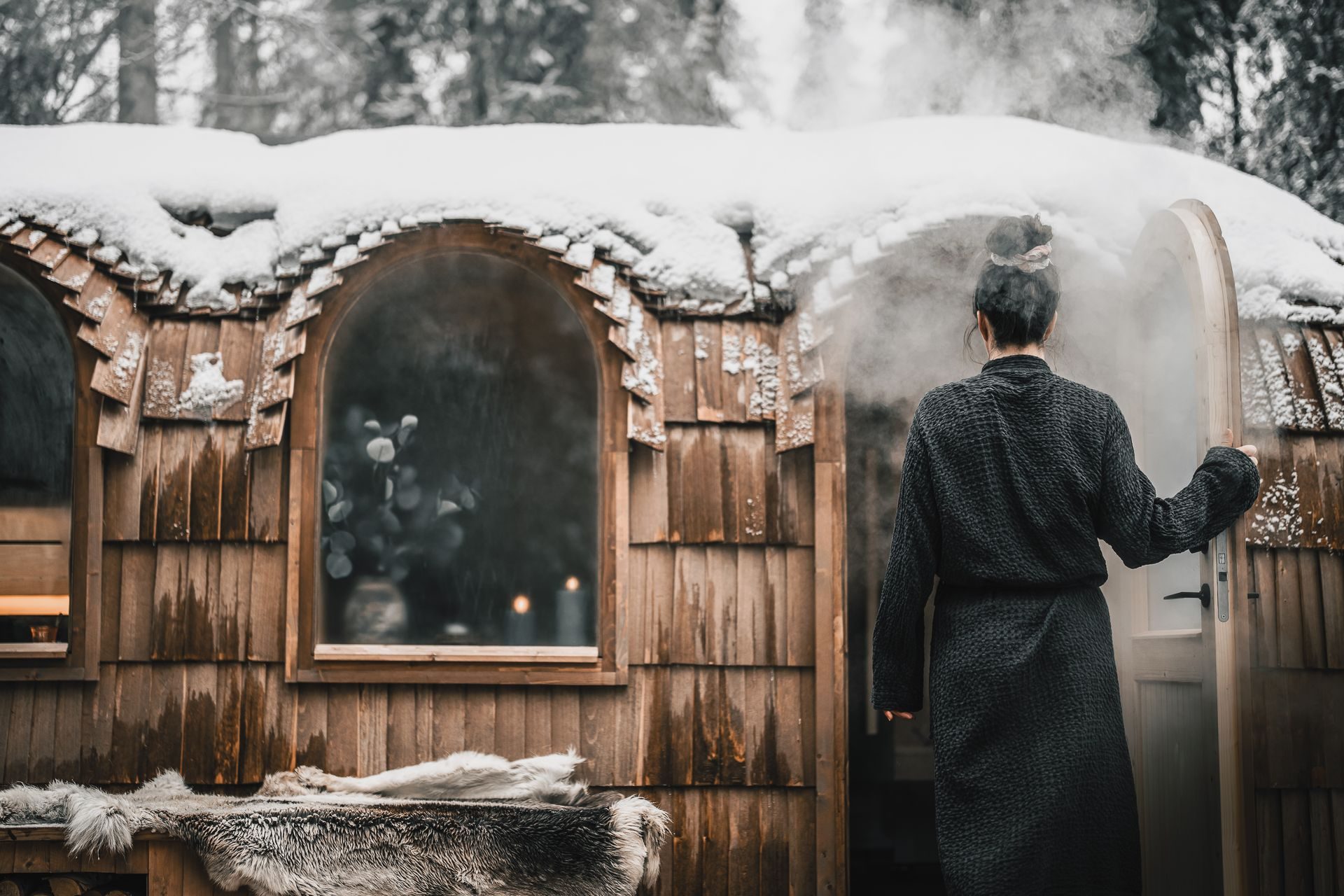 A guest entering the wooden sauna at night, snow on the timber walls.