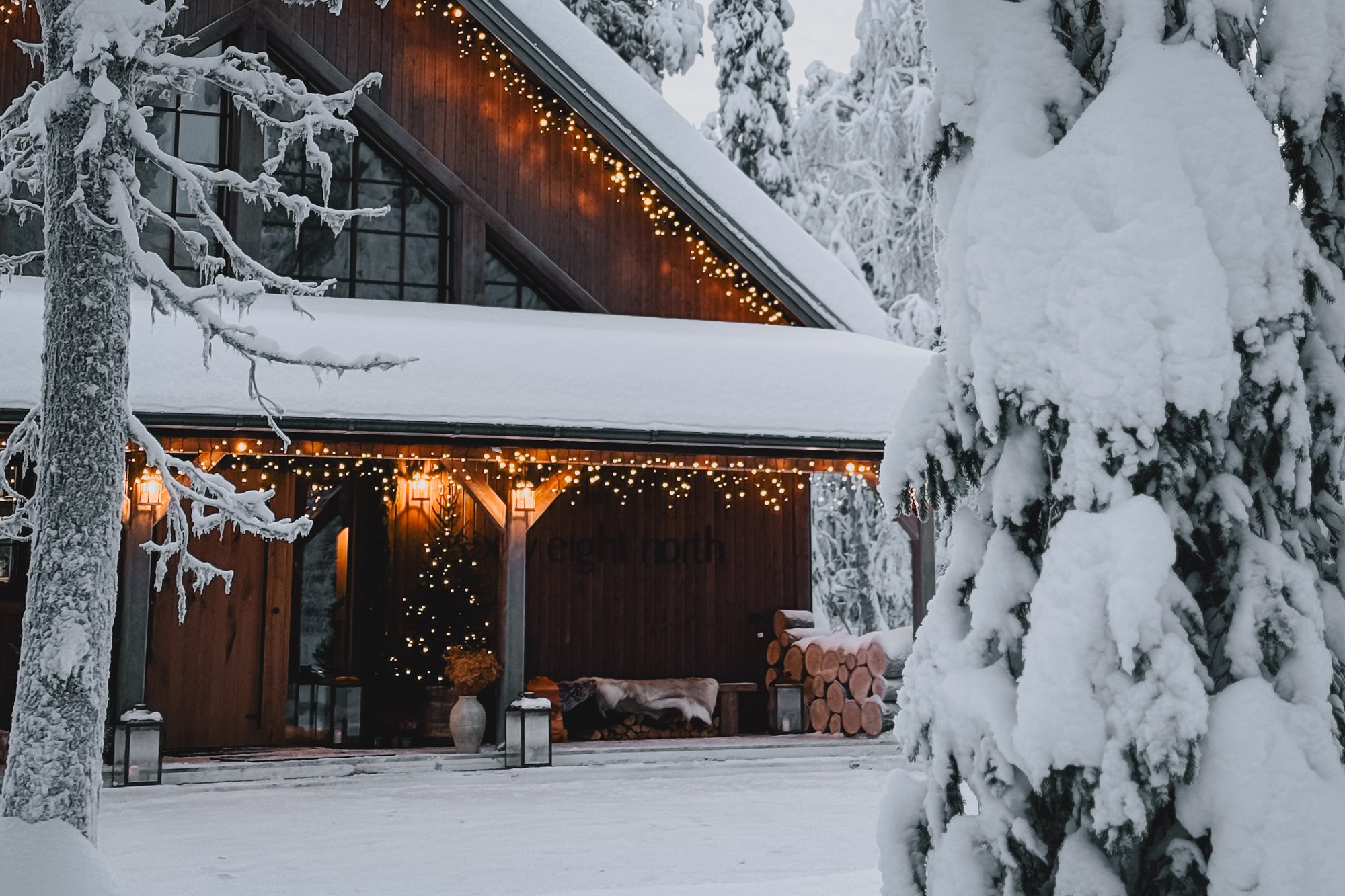 The Sixty Eight North lodge in deep winter — snow-laden pines, fairy lights along the eaves, lanterns and stacked firewood beneath the covered porch.