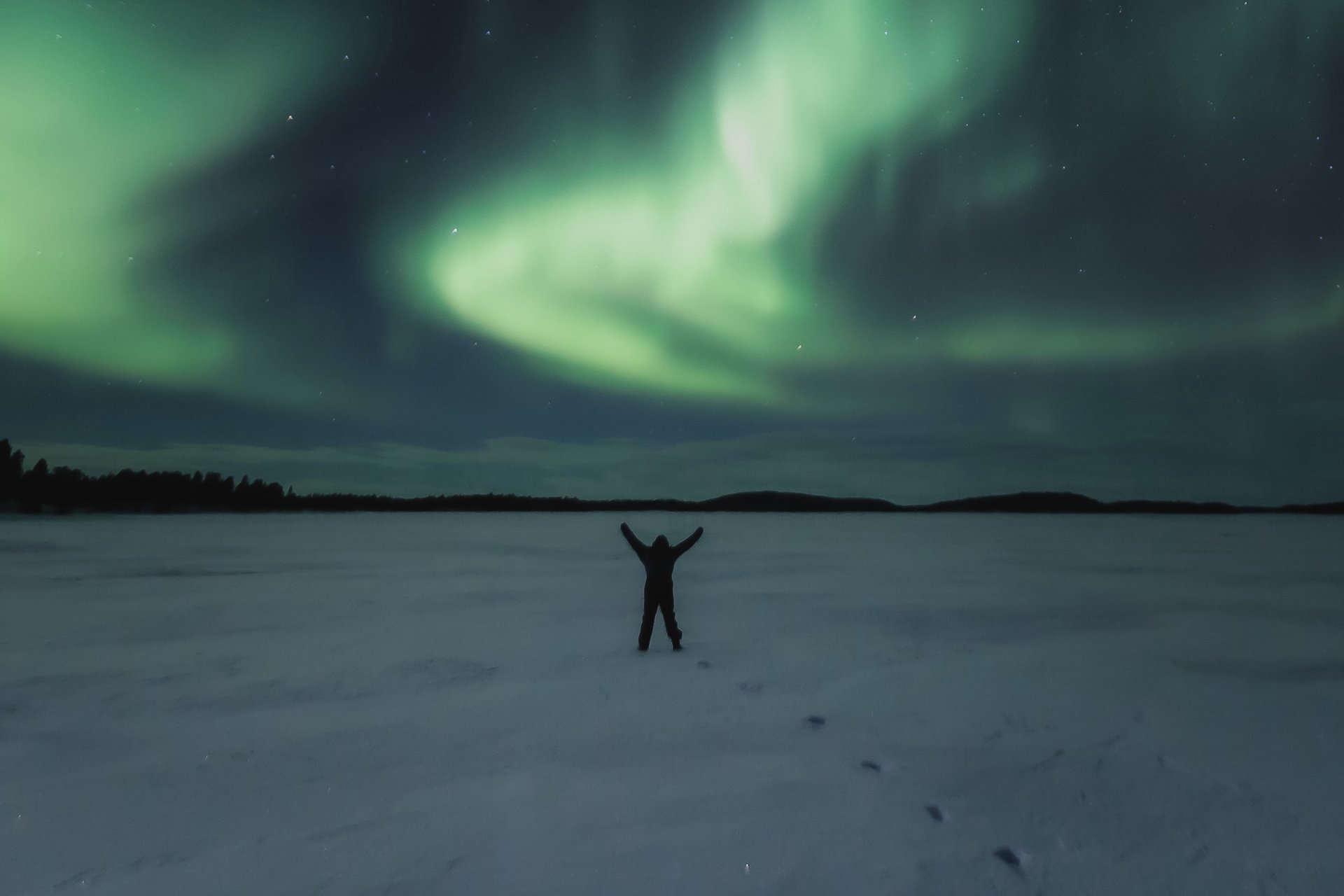 A figure on a frozen lake with arms raised under a sweeping aurora arc, the Arctic sky glowing green above.