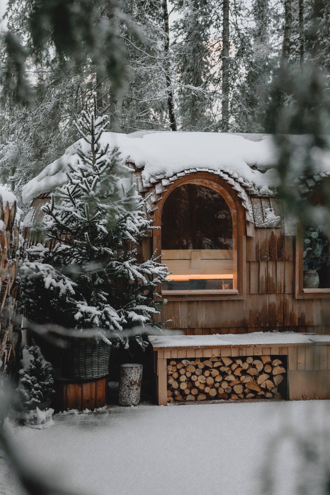 The handcrafted sauna among snow-heavy pines, the warm-lit arched window glowing from inside, firewood stacked beneath the bench.