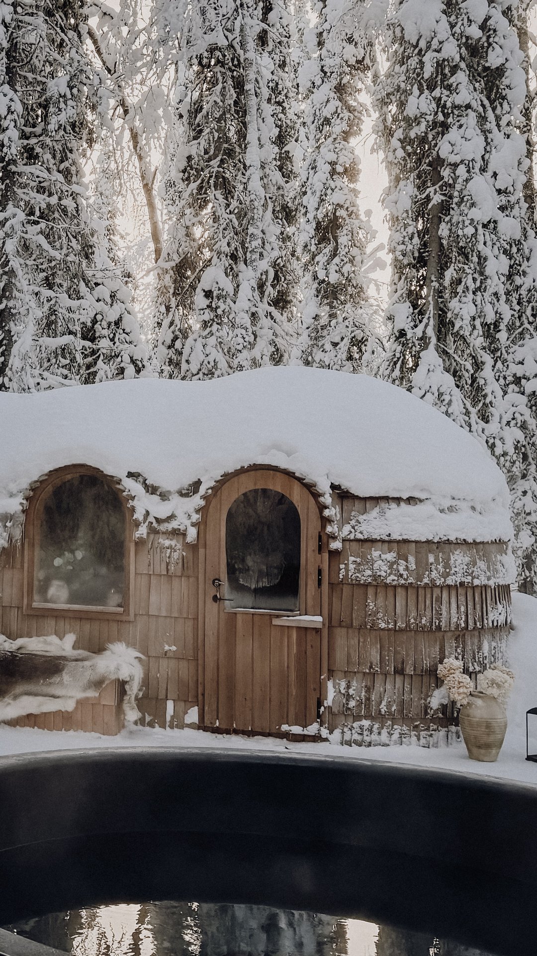 The handcrafted forest sauna with arched doorway and hand-laid shingles, snow heavy on the roof, the hot tub edge in the foreground.