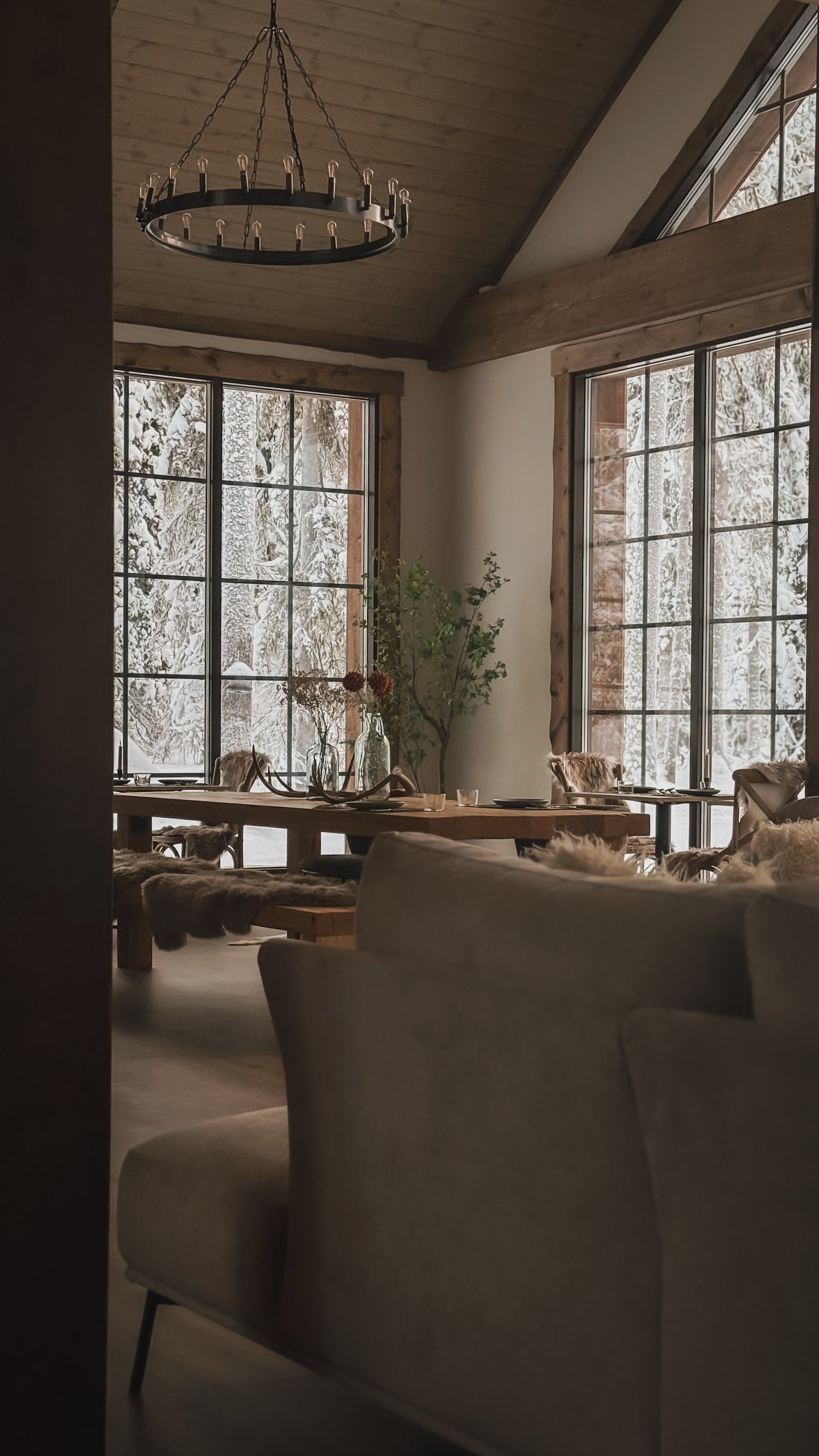 Inside the lodge at Sixty Eight North: circular ironwork chandelier above the long table set with reindeer-skin chairs, tall mullioned windows onto the snowy birch beyond.