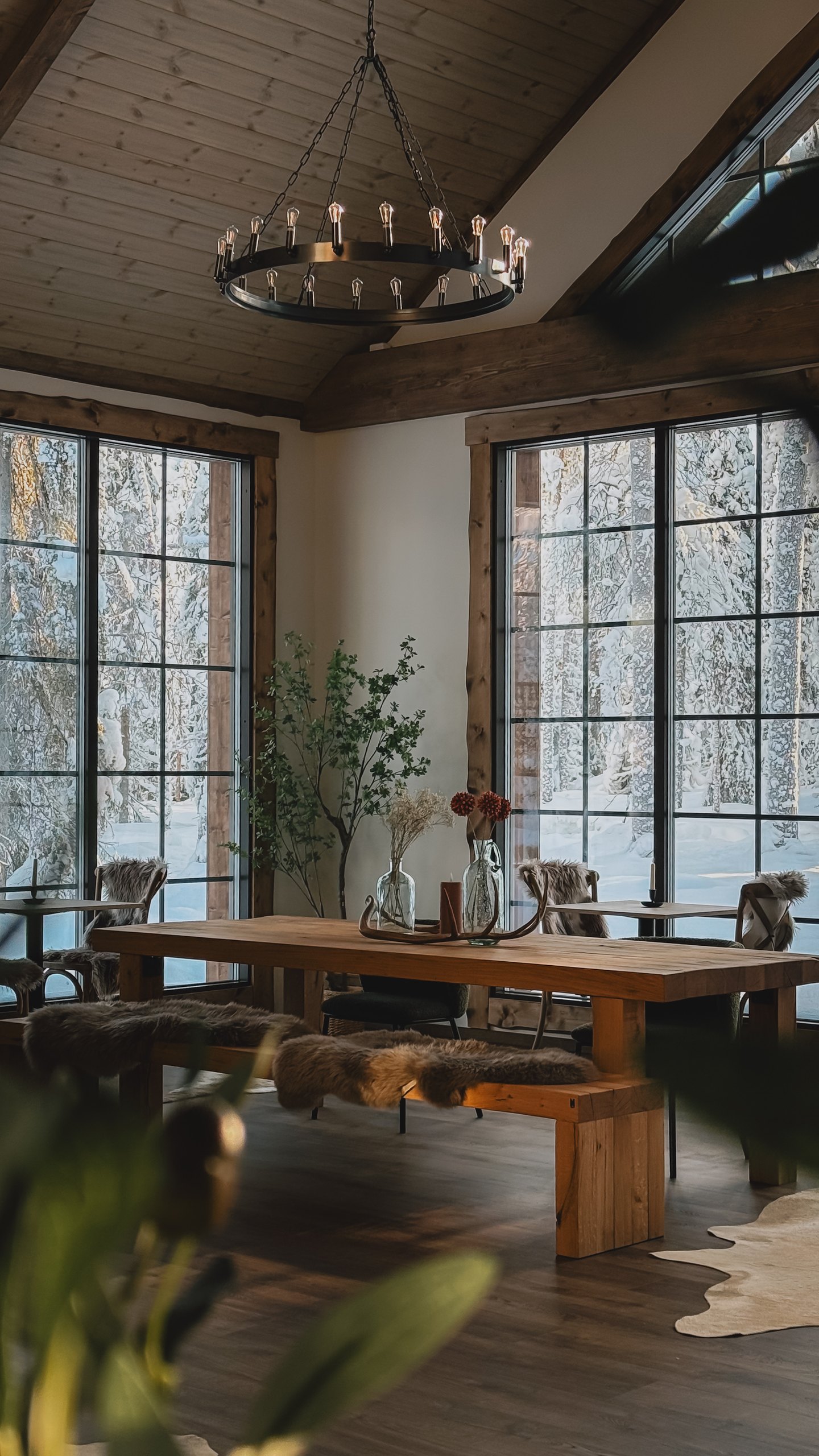 The dining room at the lodge: long wooden table, ironwork chandelier, tall windows onto snowy birch.