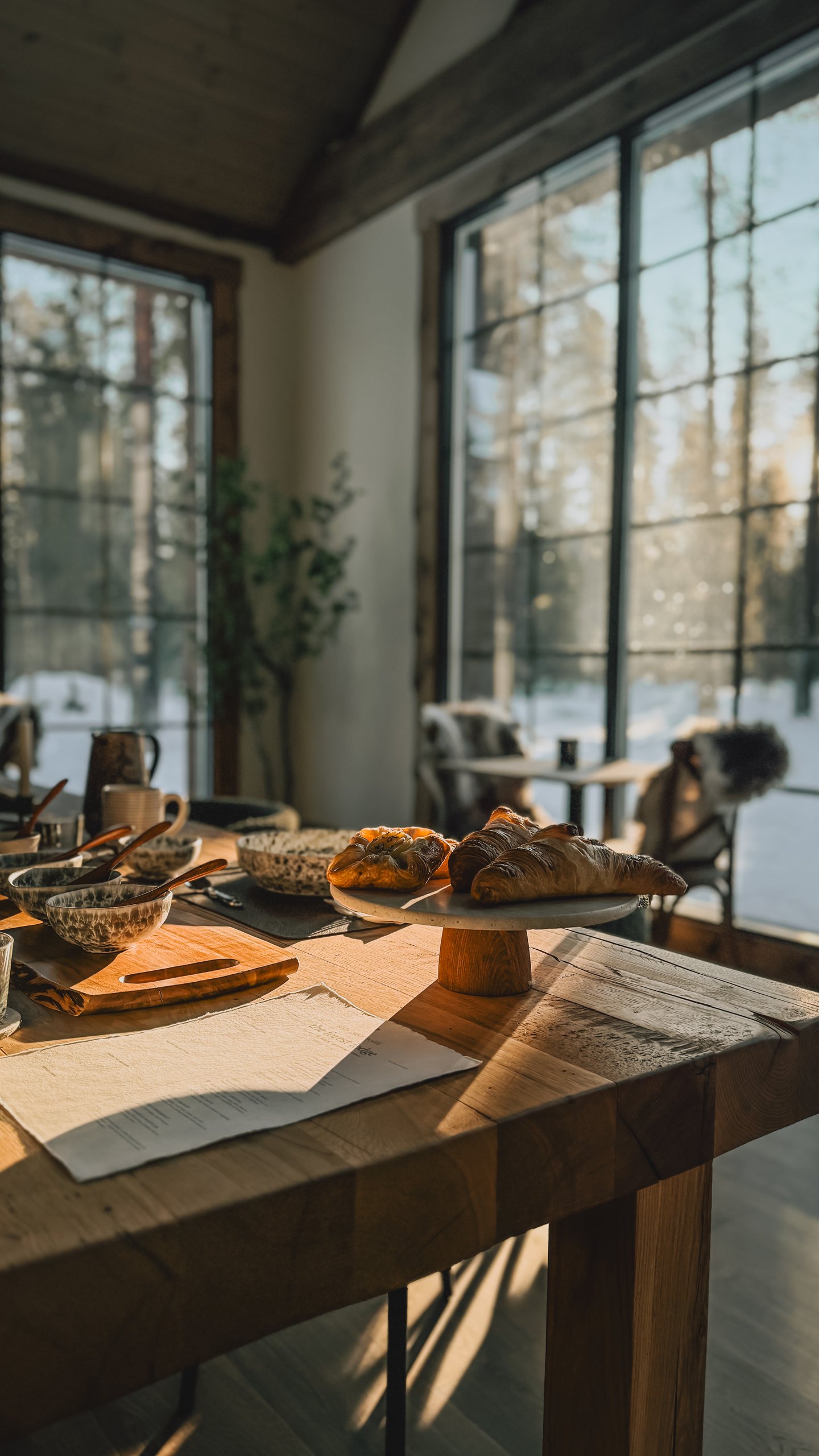 A breakfast spread on the long table at the lodge: pastries on a small stand, ceramic bowls and mugs, a printed menu, morning sunlight streaming through the tall windows onto the snowy birch beyond.