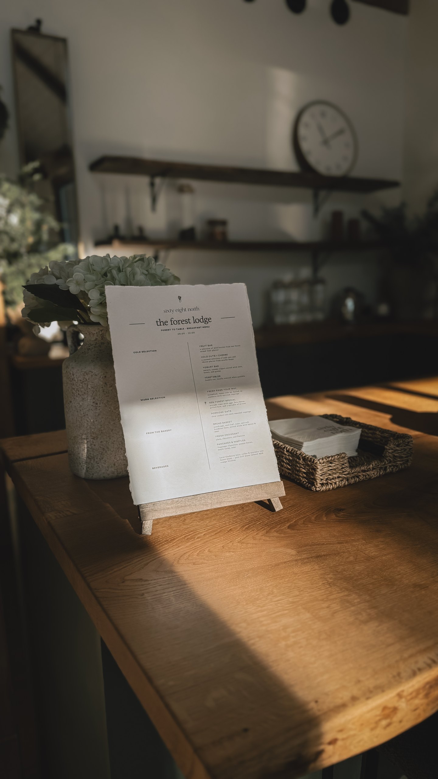 The forest lodge breakfast menu card on a small easel beside a stoneware vase of soft hydrangea, set on the wooden table in warm afternoon light.