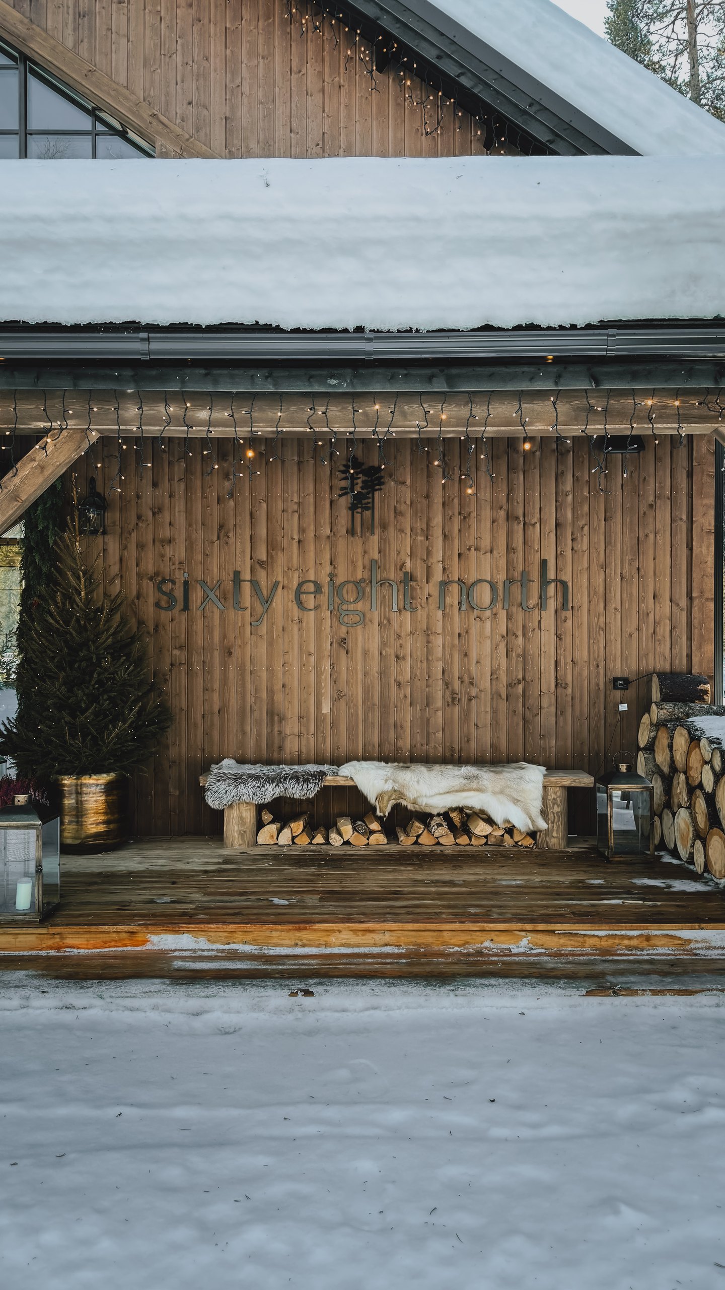 The lodge entrance at blue hour: timber wall with the Sixty Eight North wordmark, festoon lights and a small lit Christmas tree, a bench with reindeer skin over a snow-laden woodpile.