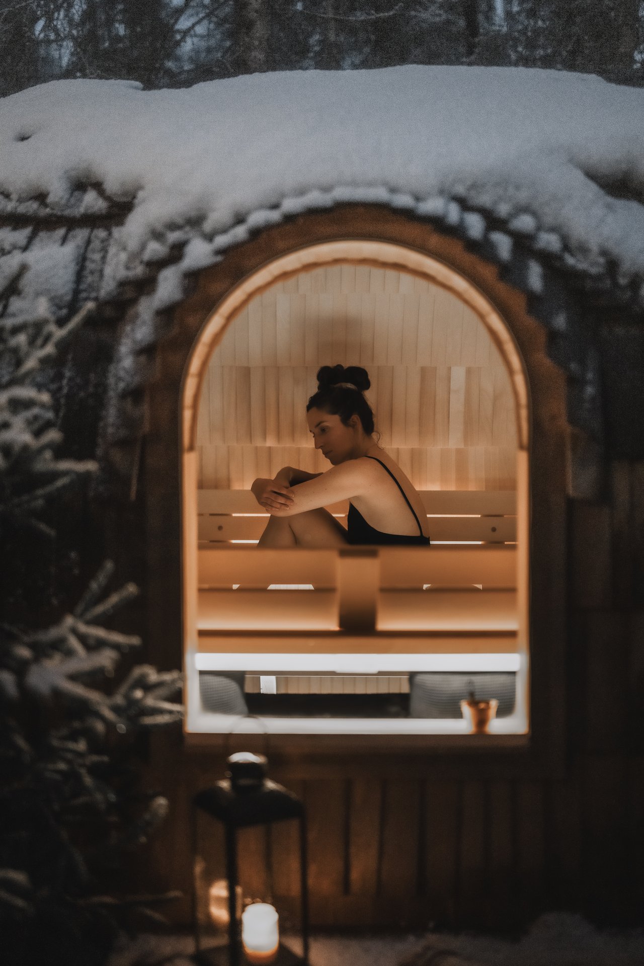 A guest seated quietly inside the snow-laden barrel sauna at dusk, the lantern lit on the path outside.