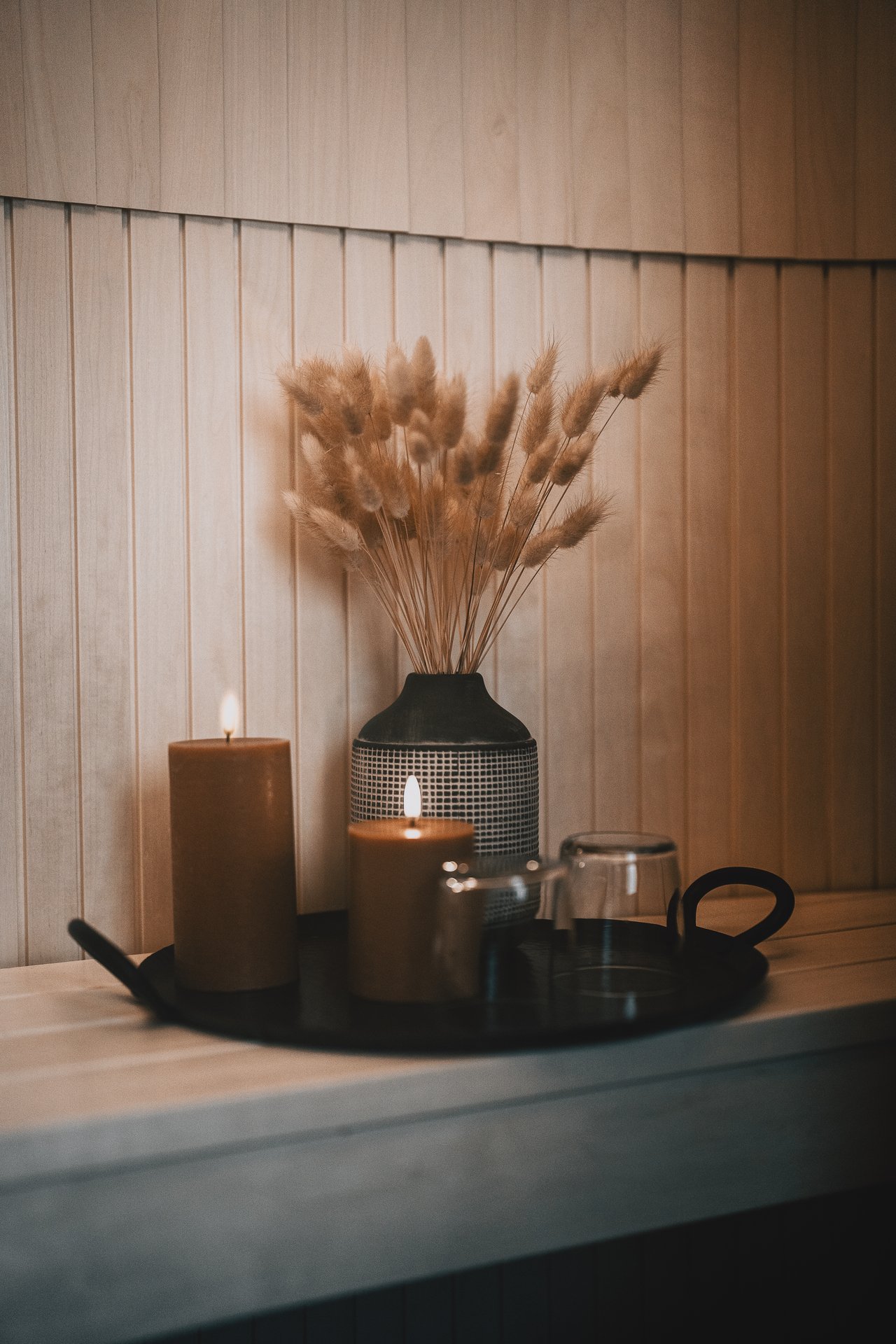 A black tray of warm-coloured candles, a striped vase with dried grasses, and a small dark glass on a wooden cabin bench: the quiet setup before a treatment.