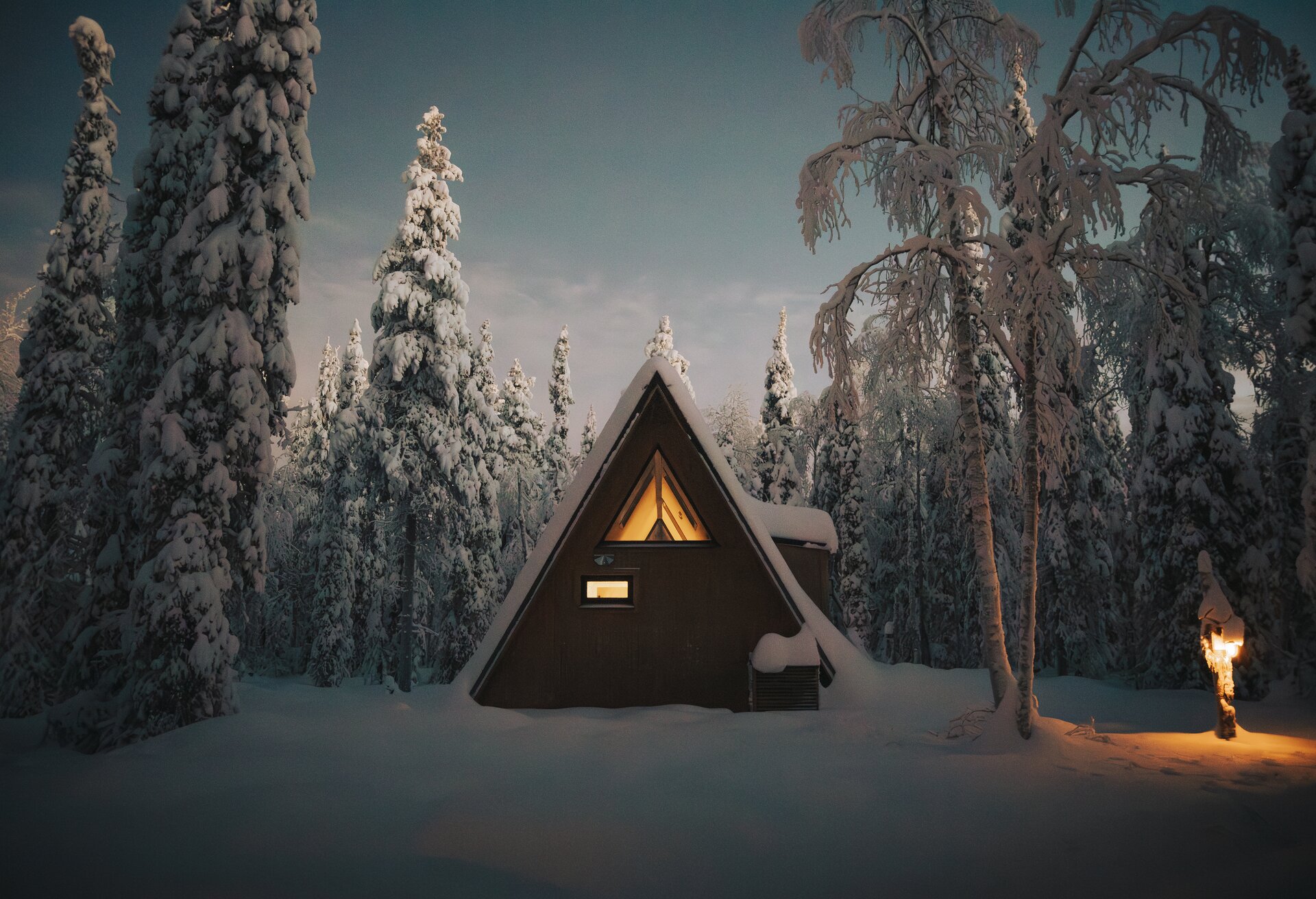 An A-frame cabin glowing warm at blue hour, snow-laden pines all around and a lantern lit on the path.