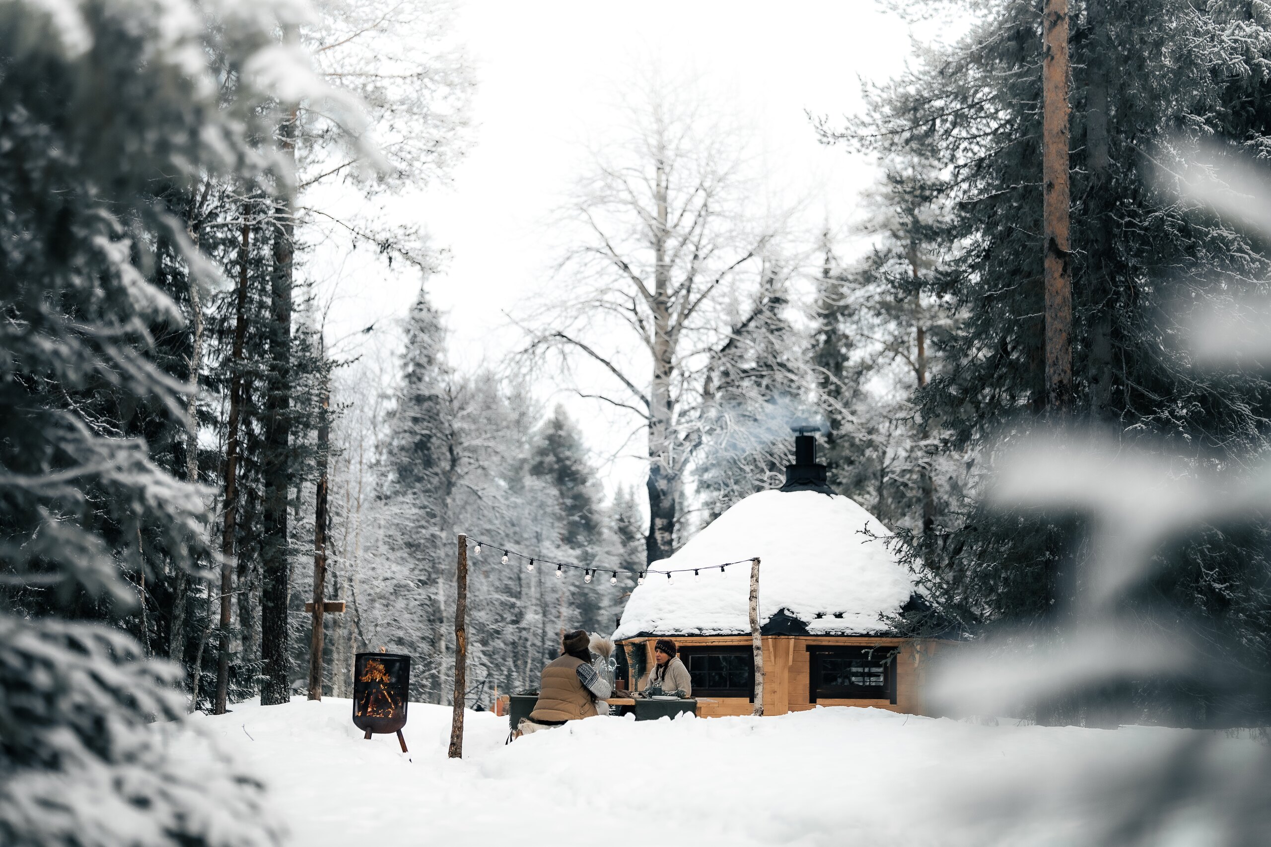 Guests gathered at a long table beside a kota in the snowy pine forest, string lights and a small fire warming the scene.