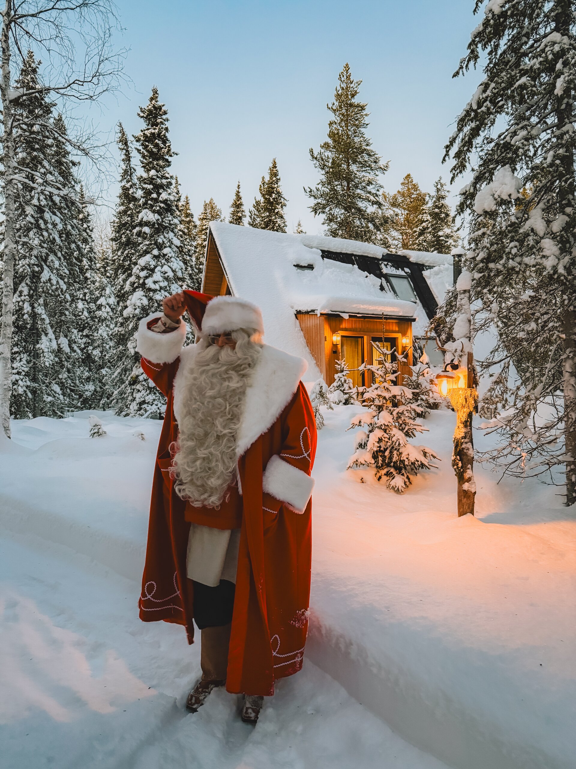 Santa walking up to a glowing A-frame cabin in the snow at blue hour, snow-laden pines and a warm-lit doorway just ahead.