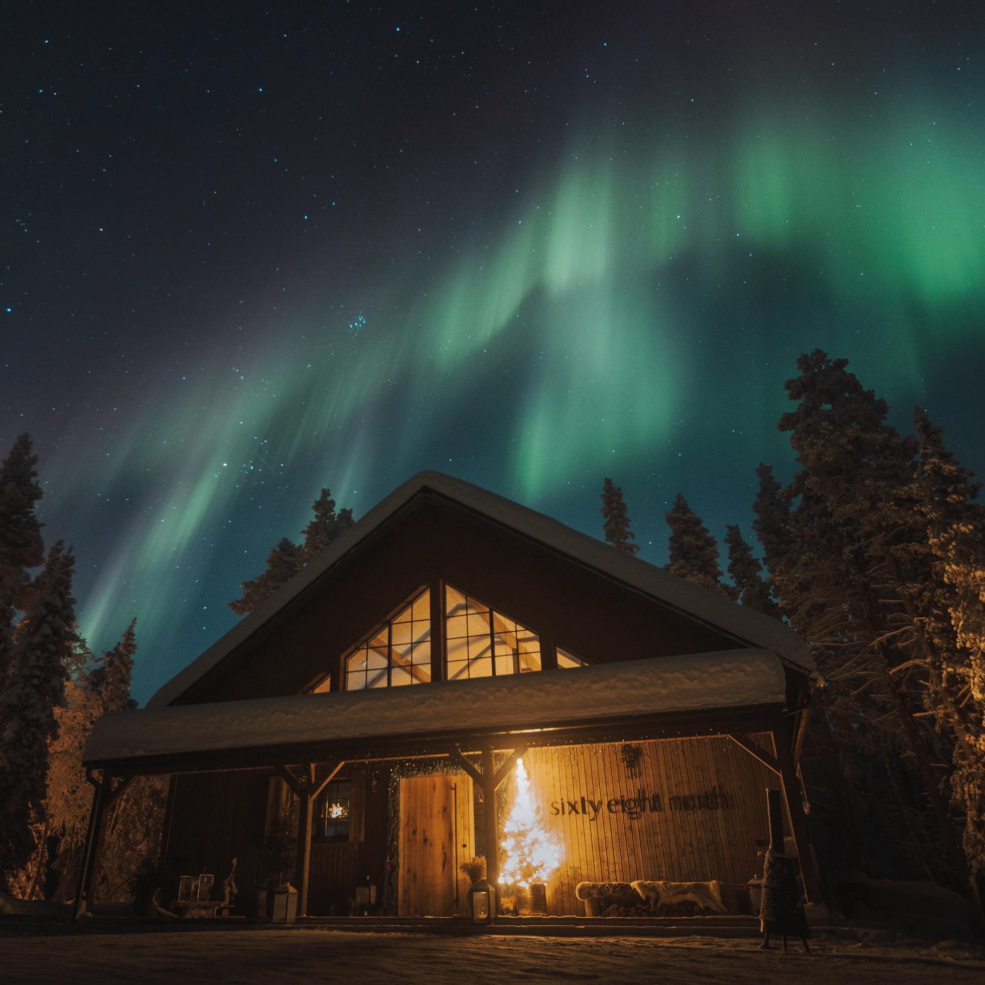 The Sixty Eight North lodge glowing warm under a sweep of green aurora, snow on the roof and arctic spruces silhouetted against the night sky.