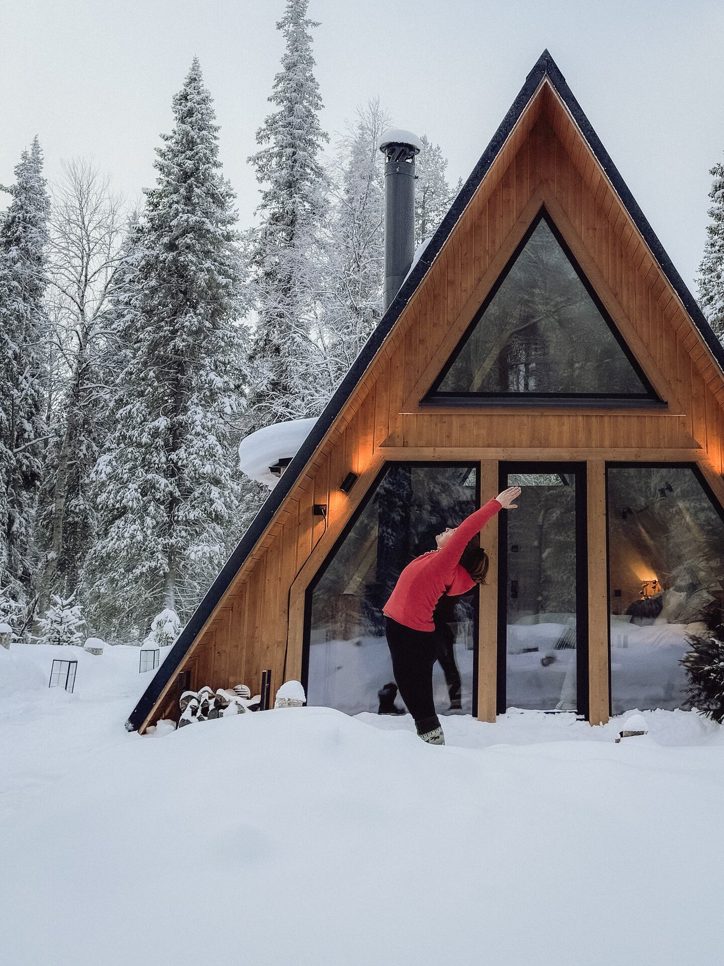 A guest in a red top in a side-stretch yoga pose in deep snow just outside the A-frame cabin, snow-laden pines all around.