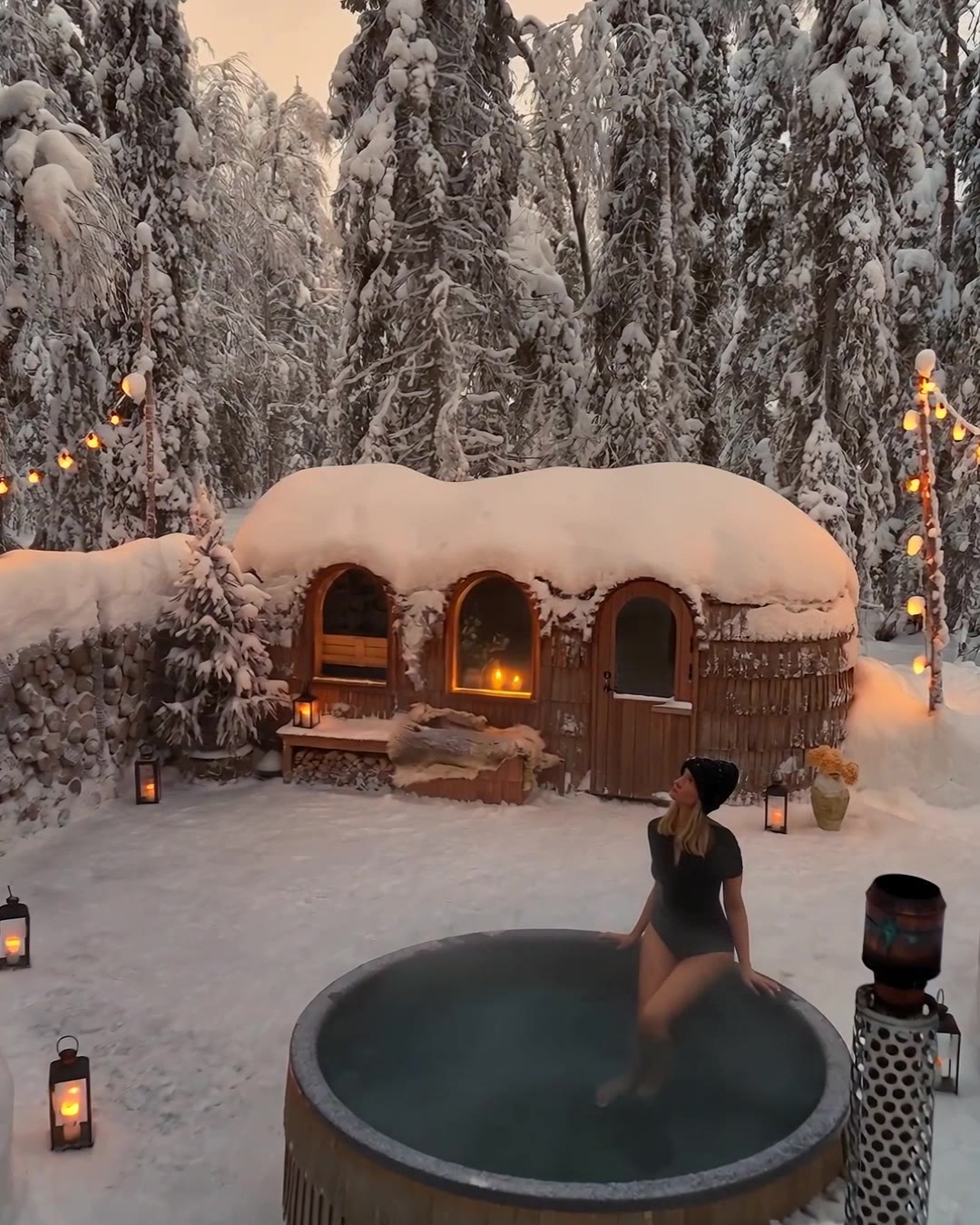 Aerial view of the forest sauna and outdoor hot tub at Sixty Eight North, the wooden sauna and stacked firewood beside, surrounded by snow-laden pines.